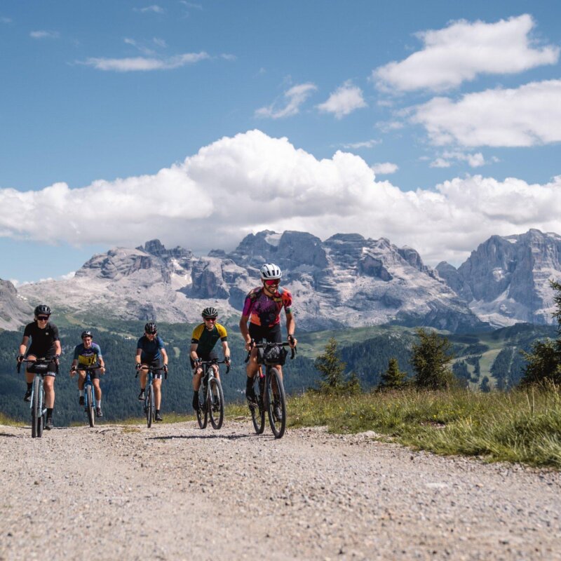 Un gruppo di ciclisti gravel pedala su una strada sterrata panoramica nelle Dolomiti, circondati da prati verdi e con maestose montagne sullo sfondo sotto un cielo azzurro con nuvole bianche. L’atmosfera è di avventura e sfida, immersi nella natura alpina.