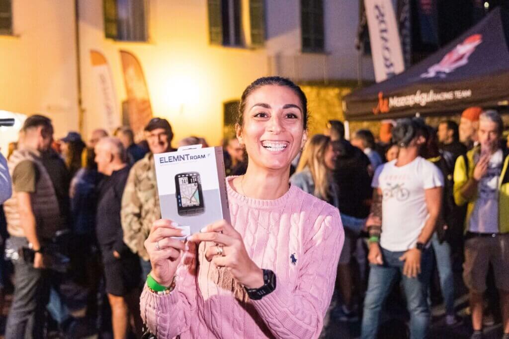Donna sorridente mostra un ciclocomputer Wahoo ELEMNT Roam appena vinto durante l’estrazione premi dell’evento Unpaved Roads Foreste Casentinesi, circondata da altri partecipanti nel village serale.